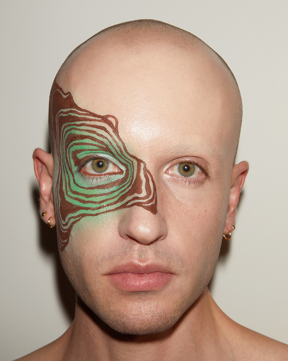 Close up portriat of Jesse at a front angle with graphic brown liner make up in concentric shapes extending out from one eye against a grey background
